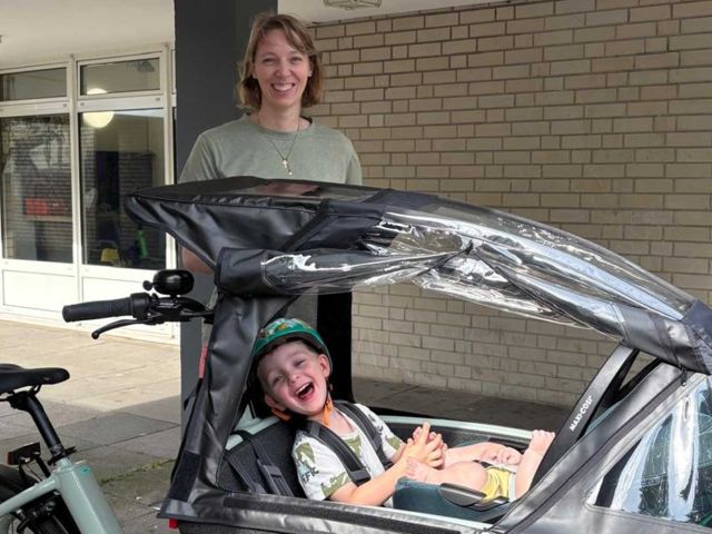Smiling woman stands beside a pedicab with a clear plastic canopy, a cheerful child sits laughing in the front seat