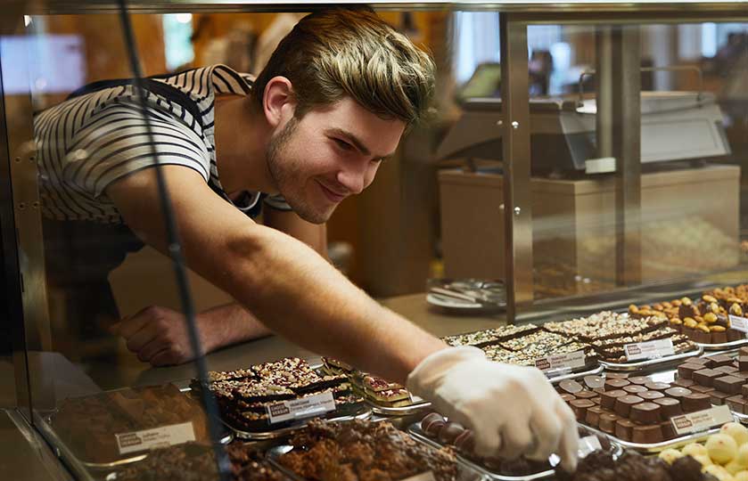 Man in a striped shirt wearing a glove reaches into a glass display case filled with assorted chocolates and sweets, selecting treats