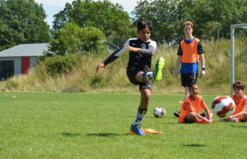 Boy in a black kit kicks a soccer ball during a youth practice, with bright blue cleats and teammates in orange bibs watching on a grassy field.