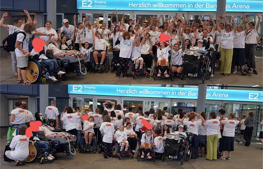 Large group photo outside an arena entrance: many people in white shirts, several in wheelchairs, raising hands in celebration; welcoming signage visible above.