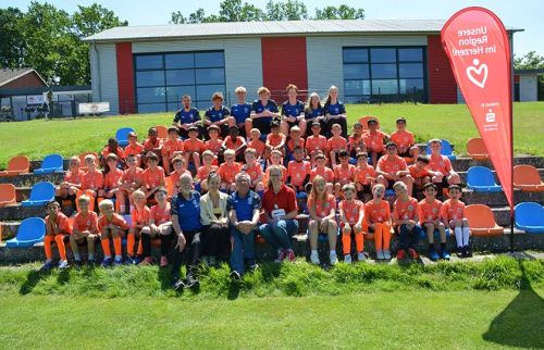 Group photo of young soccer players in orange jerseys with coaches on outdoor bleachers, a red banner to the right.