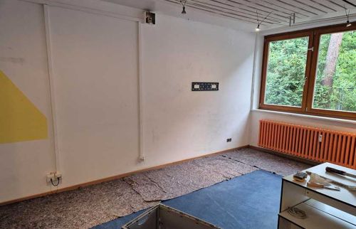 Room under renovation with bare white walls, a protective floor covering, and a large window with an orange radiator beneath.