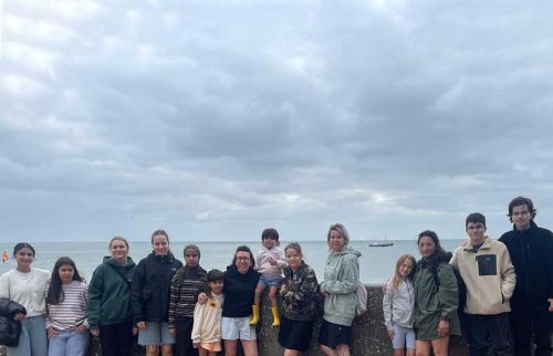 Group of people of various ages posing together along a sea‑front wall on a cloudy day, with the ocean and a boat in the distance.