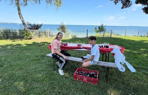 Two children sit at a lakeside picnic table with a red tablecloth, drinks and snacks, and a crate of bottles nearby on the grass.