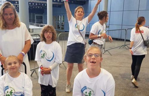 Group of smiling children and adults in white event shirts pose outside a building, some with arms raised in celebration.