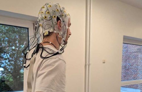 Side profile of a person wearing an EEG cap with multiple electrode wires attached indoors.