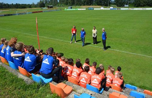 Group of young players in blue and orange jerseys seated on colorful stadium seats, listening to four coaches on a grassy soccer field nearby on a sunny day.