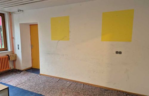 Empty room under renovation with two yellow wall patches and a yellow door, blue carpet, and a window with a radiator nearby.