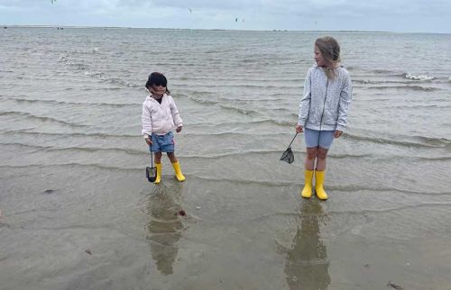 Two children stand in shallow beach water wearing yellow rain boots, each holding a small net, with the ocean and distant kite surfers in the background.