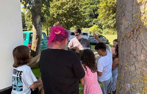 Group of adults and children gathered around a table outdoors, likely at a community event in a sunny park.