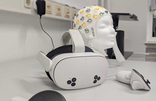 White VR headset with strap on a lab bench, alongside a detachable controller and a mannequin head wearing a patterned cap in the background.
