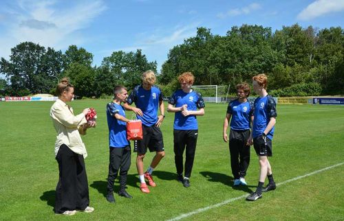 A woman presents a red gift bag to a young soccer player on a sunny green field, teammates watching nearby.