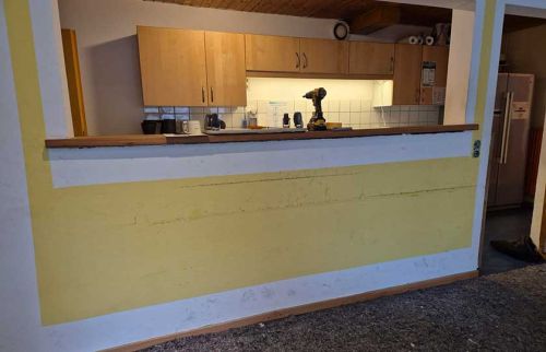 Open kitchen pass-through with wooden cabinets, tiled backsplash, and a drill on the counter visible through the opening in a yellow-walled room.