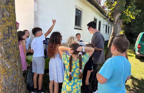 Group of kids gathered around a table outside a white building as an adult hands out papers or items to them.