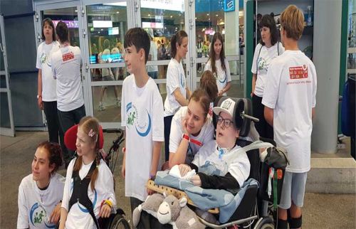 Group of young volunteers in white shirts at a charity event, with two children in wheelchairs in front and others standing nearby near glass doors.
