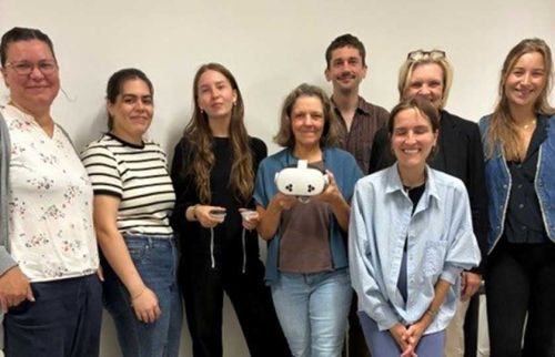 Group of nine adults posing together in an office, with the person in the center holding a white VR headset or similar device.
