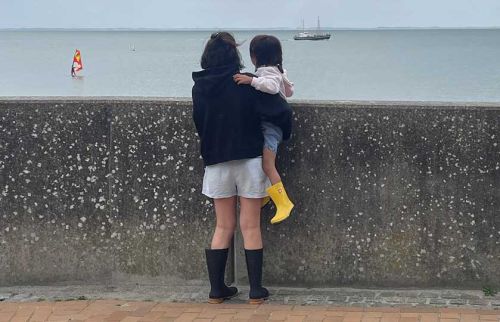 Two people stand at a seawall, an adult with a child facing the water, the adult wearing a dark hoodie and white shorts while the child has yellow rain boots and a light pink top near the ocean.