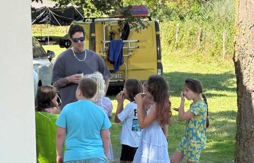 Man wearing sunglasses talks to a group of children outdoors near a yellow van, he holds a cup.
