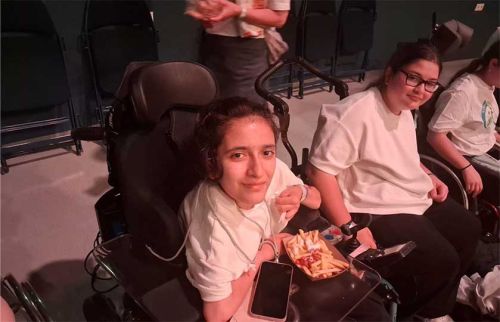Two young women in wheelchairs at an event; the front girl holds fries and a phone, looking at the camera.
