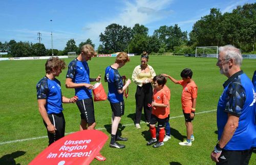 Youth soccer players in blue and orange jerseys receive awards on a sunny grass field as coaches hand out prizes. A banner with German text is visible in the foreground.