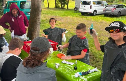 Children in plastic ponchos at a park table slicing watermelon under supervision while a man watches; vans parked in the background.