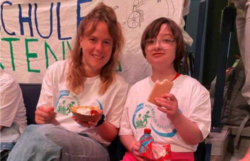 Two smiling people in white shirts sit on a bench, eating snacks; one holds a bowl, the other a wafer and a juice box, with a banner in the background.