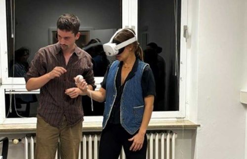 Woman wearing a white VR headset stands next to a man who is guiding her while they handle a small object in a bright room. This describes the VR activity and collaborative setup without identifying individuals.