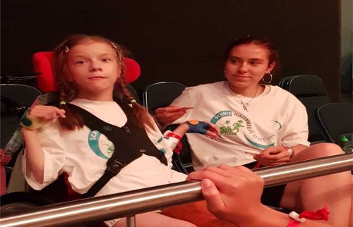 Young girl in a wheelchair wearing a white shirt and harness, with a female coach guiding her at a gym bar area.
