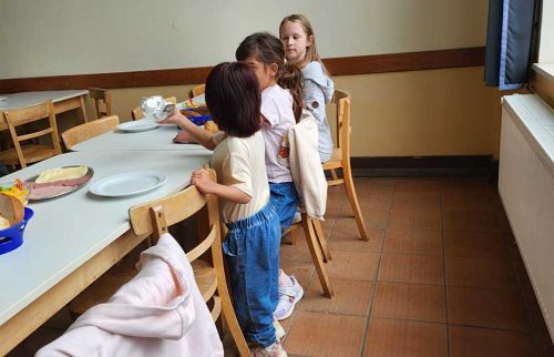 Three girls stand at a long dining table in a school cafeteria, waiting with plates and food trays nearby.