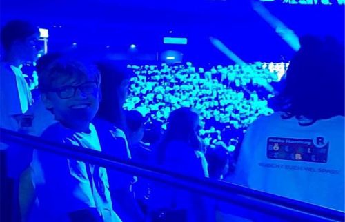 Smiling person with short hair and glasses standing on a balcony, blue lighting, crowded arena behind them.