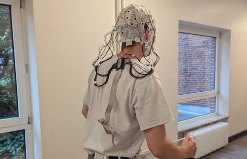 Person wearing an EEG cap with dozens of wires and electrodes, standing in a bright room.