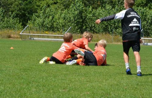Three young soccer players in orange shirts sit and lie on a grassy field during practice, as another player stands nearby.