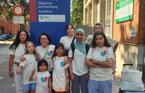 Group of diverse women and children in matching white shirts with a heart logo pose together outside a building.