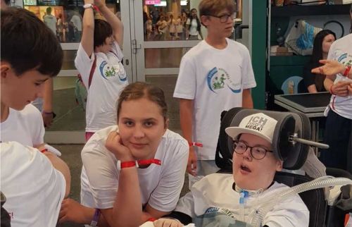 Teen girl in a white shirt rests her chin on her hand beside a person in a motorized wheelchair with breathing equipment at a charity event with volunteers nearby.
