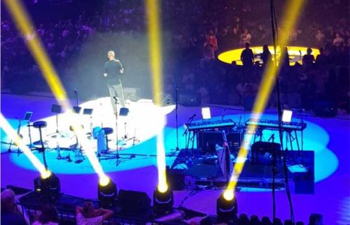 A performer stands on a lit circular stage with keyboards and stools, yellow spotlights streaming down into a seated audience.