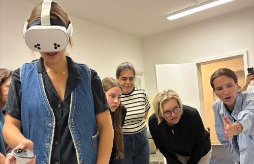 Person wearing a virtual reality headset and denim vest, holding a controller as a group watches a demonstration in a classroom.