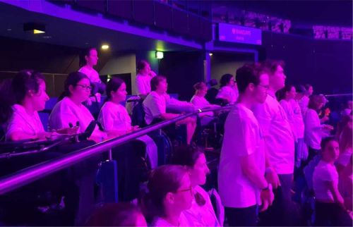 Group of people standing and watching an event in a dimly lit arena with purple lighting along a railing.