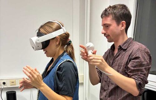 Woman wearing a VR headset while a man assists with a white handheld device in a bright lab setting.