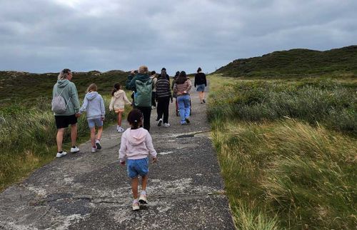 Group of people, including children, walking along a cracked path through grassy dunes under a cloudy sky.