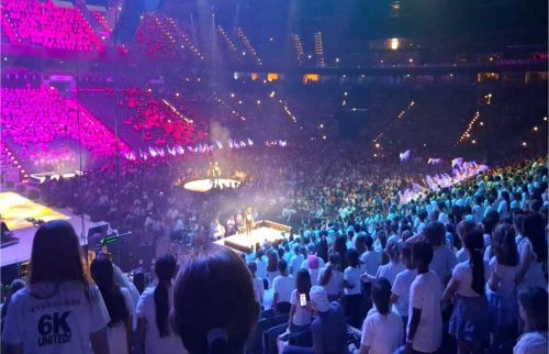 Large indoor arena filled with a crowd watching a concert; purple stage lights and performers on a circular platform.