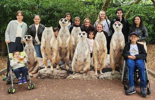 Group of smiling people posing outdoors with a row of standing meerkats at a park, including a child in a stroller and a person in a wheelchair.